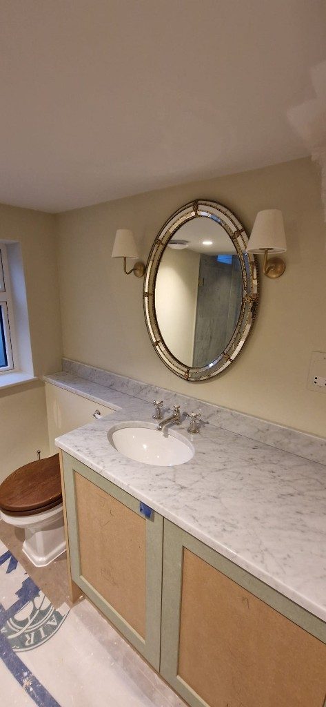 Bathroom vanity install with marble top, oval gold mirror and brass wall lights by Ikveles Construction